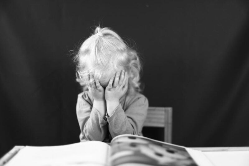 How Movement and Early Intervention Can Support Children With Dyslexia 1 Black and white photo of a young child covering their face with hands, sitting in front of a book.